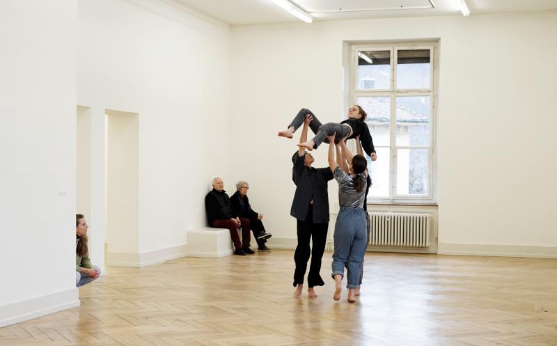 Drei Tänzerinnen halten eine vierte Tänzerin mit ausgestreckten Armen hoch in die Luft. Hinter ihnen sieht man ein älteres Paar, welches auf einer weissen Bank sitzt, sowie eine junge Frau, die im Türrahmen kauert, gebannt zuschauen.