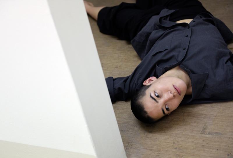 Eine Tänzerin, die auf dem Rücken am Boden in einem engen Raum liegt und mit geneigtem Kopf durch die Türöffnung direkt in die Kamera blickt.
