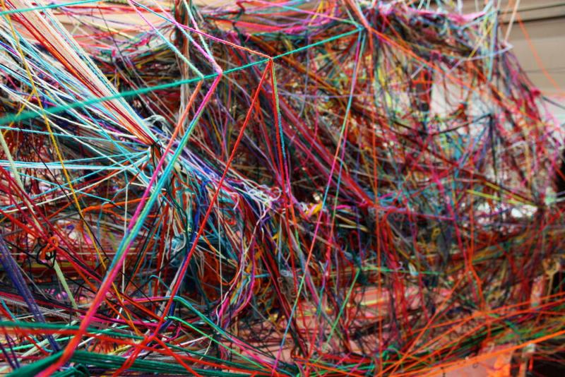 a dense, unpatterned structure made from many different and colourful wool strings. The wool runs diagonally and crisscrosses chaotically across and ties to a geometric-like structure under the wool.  