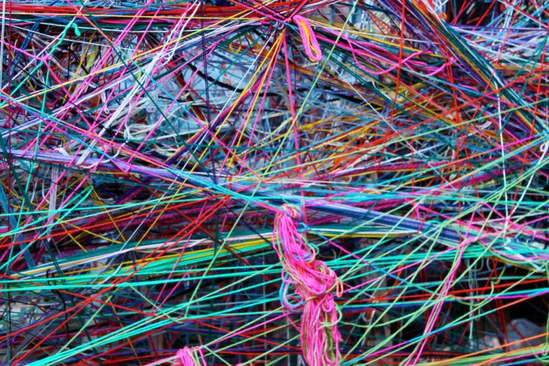 a dense, unpatterned wall structure made from many different and colourful wool strings. The wool runs diagonally and crisscrosses chaotically across the image. There is a pink, messy knot of woll lower middle of the image. 