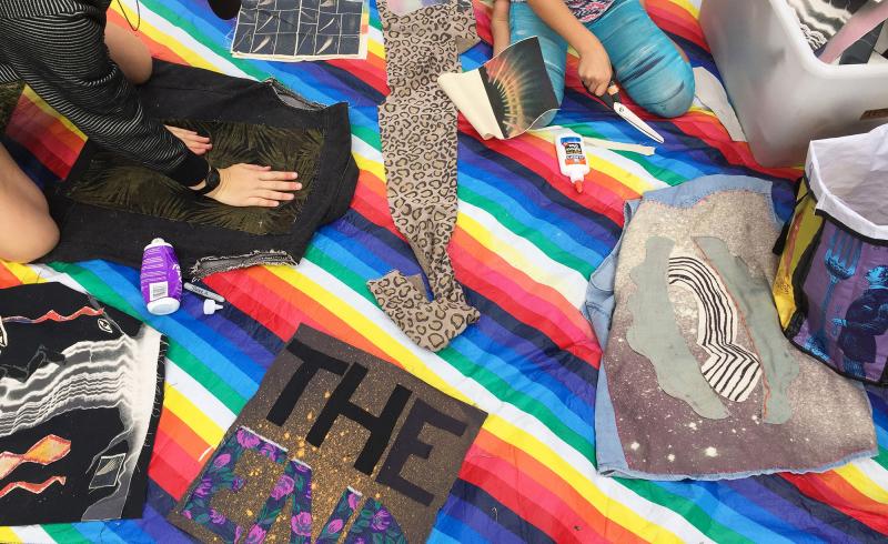 Documentation of a textile-based workshop. Several people of different ages are sitting on a rainbow blanket. They are using glue and scissors to make appliqué art on fabric and clothing.