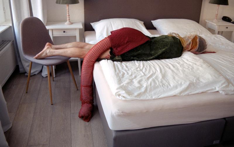 Zu sehen ist aus der fotografische Serie "In H." aus dem Jahr 2022, bestehend aus 15 Motiven, ein Einzelbild aus der Reihe. Es zeigt eine Person im Kostüm halb auf einem Bett / halb auf einem Stuhl liegend. 