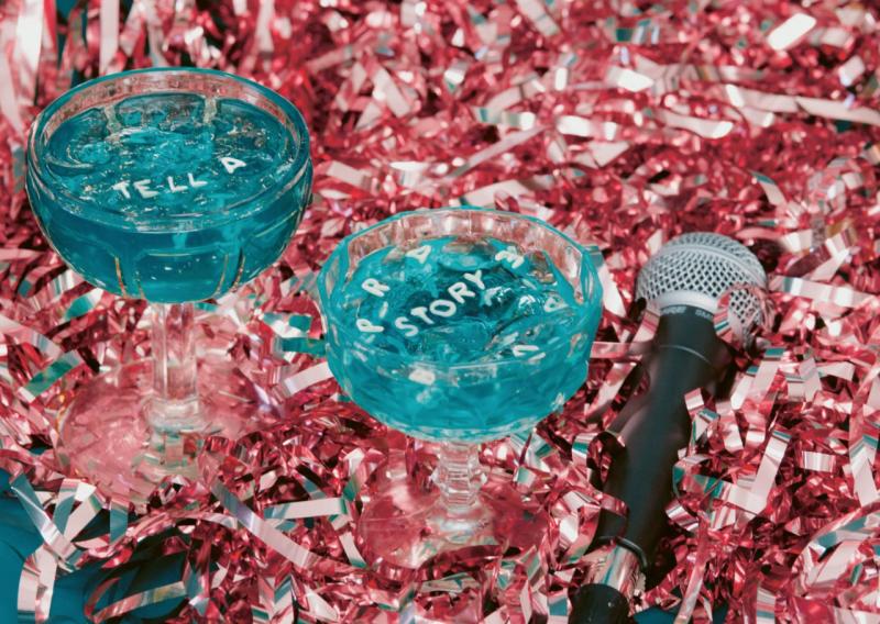 Im Hintergrund Glitzer-Lametta und ein Mikrofon, im Vordergrund zwei Cocktail-Gläser mit blauer Flüssigkeit, darin schwimmen Buchstaben, die die Worte "Tell a Story" bilden