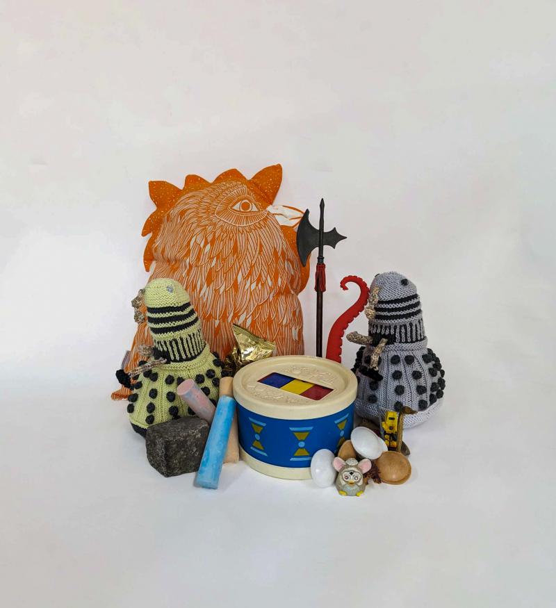 Collection of toys and objects displayed on a grey studio background. Consisting of (from the middle going clockwise) an orange chicken shaped pillow, toy axe, an octopus tenticle, crochet robot, lego man, brass swan ornament, spaceship shaped candy, small toy dinosaur, small furby keychain, childrens toy drum, fortune cookie in its wrapper, three sticks of coloured chalk, a cube shaped rock, a crochet robot