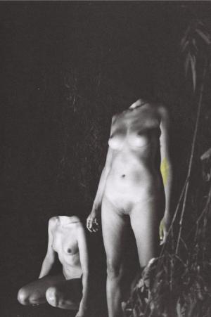 Fotografie auf der zwei nackte Frauenkörper ohne Kopf vor dunklem Hintergrund zu sehen sind.