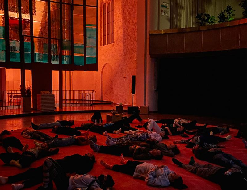 Neben einer lichtdurchlässigen Installation am linken Bildrand liegen mehrere Menschen bei gedimmtem rötlichen Licht auf einem Teppich entspannt auf dem Boden.