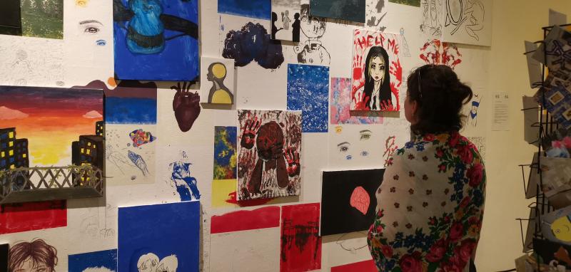 Eine Frau steht vor einer Wand. Die Wand ist mit vielen Bildern tapeziert und behängt ist. Die Frau schaut sich die Werke der Kinder an. 