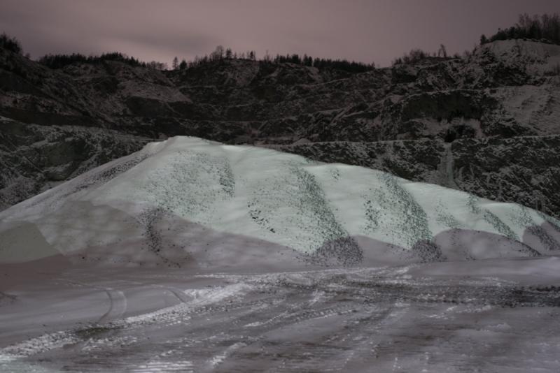 Die Fotografie zeigt einen Schutthaufen, der mit Schnee bedeckt ist, bei Nacht. Kennzeichnend sind die Farben schwarz, weiß und lila. Die lilane Farbe reflektiert am Fuße dieses kleinen Berges. Sie ist nicht künstlich erzeugt, sondern deutet auf die Lichtverschmutzung hin, die an diesem Ort herrscht.