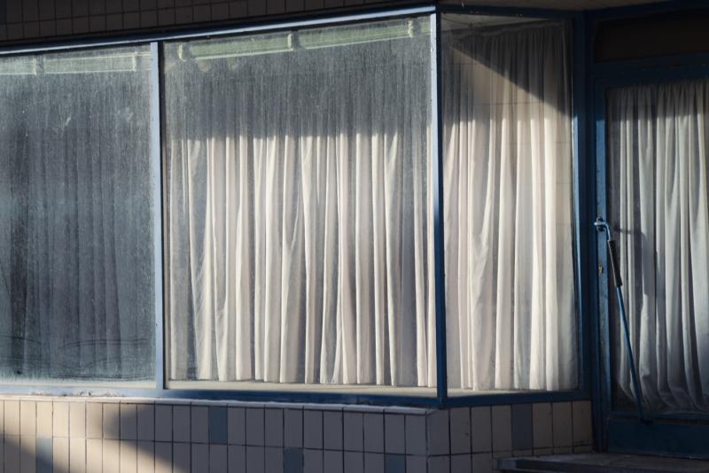 Zu sehen ist ein Schaufenster, durch das die Mittagssonne scheint. Es ist leer, bis auf einen weißen Vorhang, der das Innenleben verdeckt.