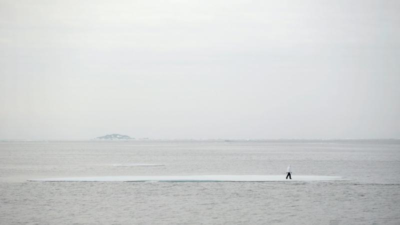 Abgebildet ist eine Eisscholle auf dem Meer, auf der eine Person läuft. Farblich hält sich alles in hellen Grautönen. Das Bild ist ein Videostill und in der Arktis gedreht.