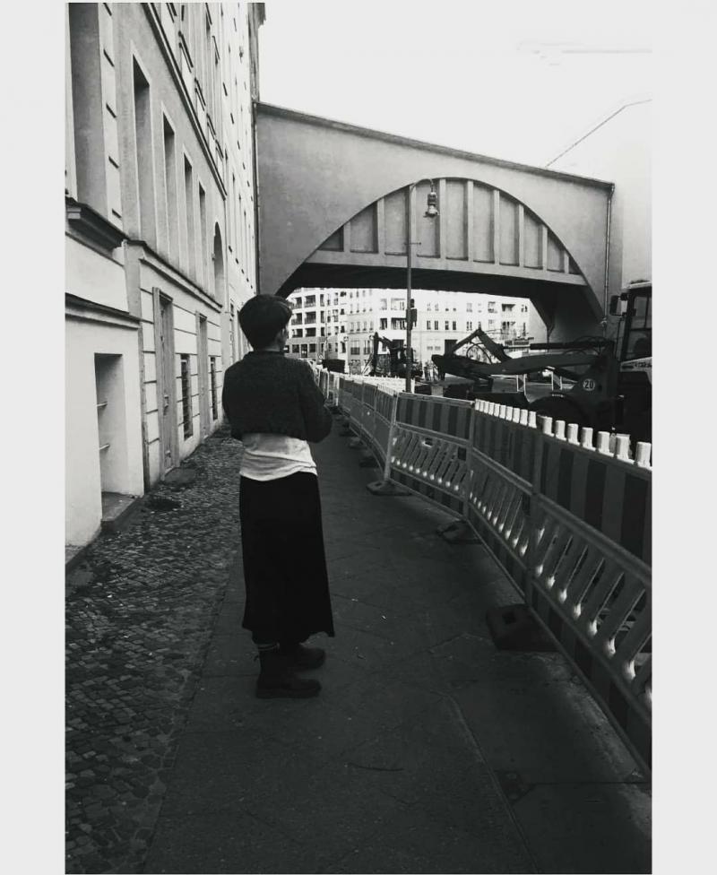 The black and white photograph presents a woman standing alone in a street. The backround is an urban space, with banners, trucks and constructions made on the streets.  