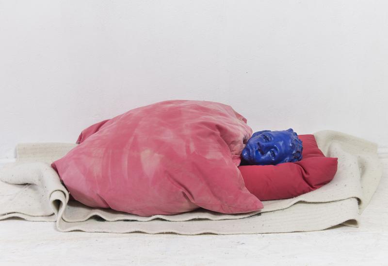 Der skulpturale Kopf eines Menschen, der auf dem Boden liegt, zugedeckt mit einer roten Decke.