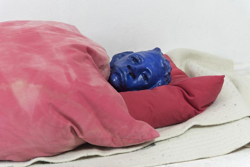 Ein blauer skulpturaler Kopf eines Menschen, der unter einer roten Decke auf dem Boden liegt.