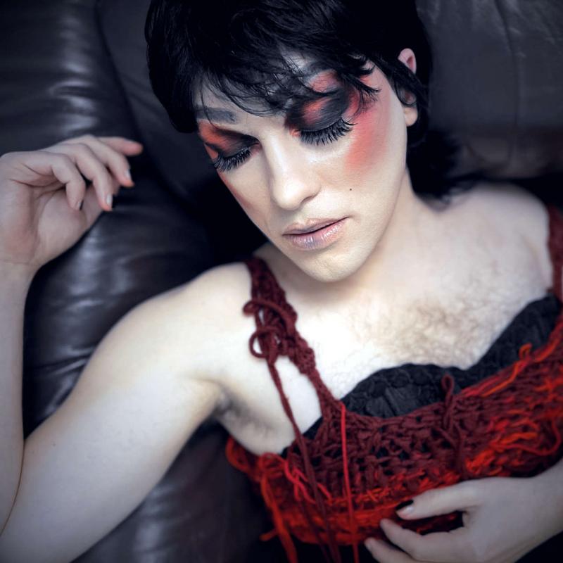 Die Drag Persona Becky liegt träumerisch auf einem schwarzen Ledersofa, nur ihr Oberkörper, ihr rechter Arm und ihr Kopf sind zu sehen. Ihre Augen sind geschlossen. Das rot schwarze Make-up leuchtet genauso wie ihr Oberteil in den gleichen Farben. 