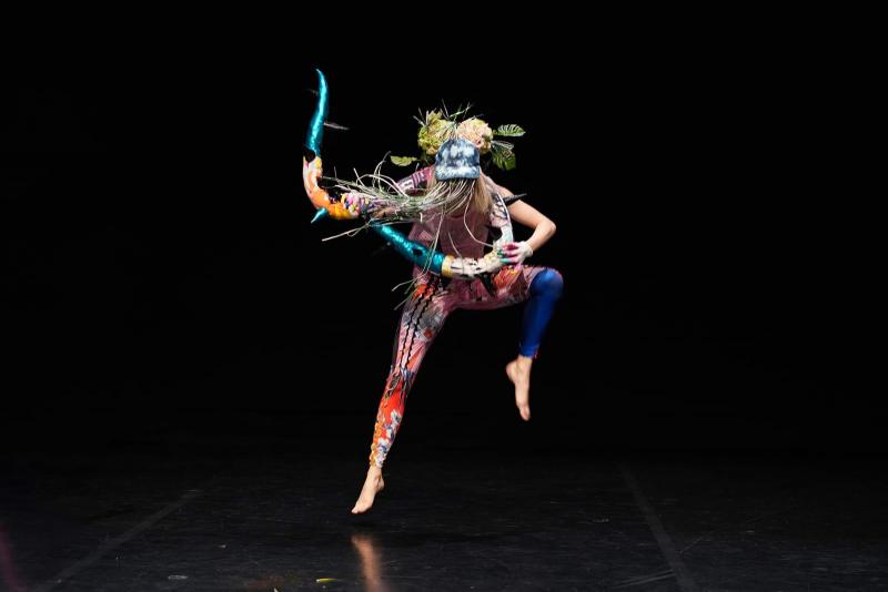 Luna Weis in the piece “Unkraut” choreographed by Doris Uhlich. She is dancing wearing colourful clothes and a mask with several elements upon it, like plants etc. 