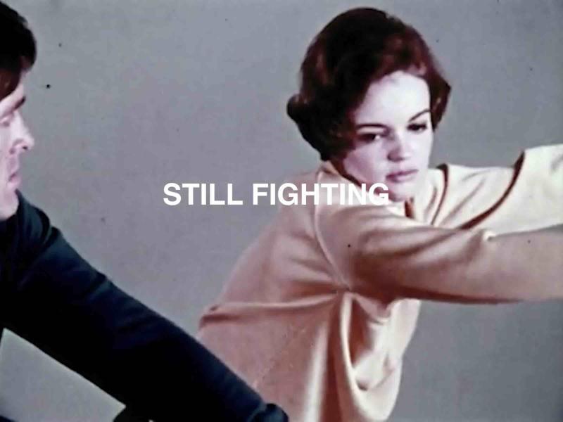 A person read as a woman on the right side and a person read as man on the left corner. A gray background. In the middle the title: "STILL FIGHTING"