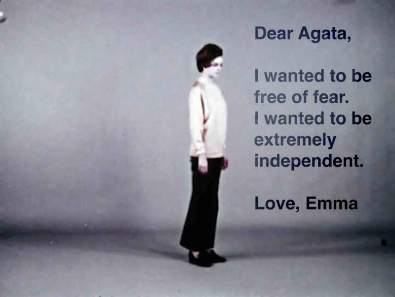 A person read as a woman, light colored against the grey background. She stands in the middle of the scene. A text that says: "Dear Agata, I wanted to be free of fear. I wanted to be extremely independent. Love, Emma" 