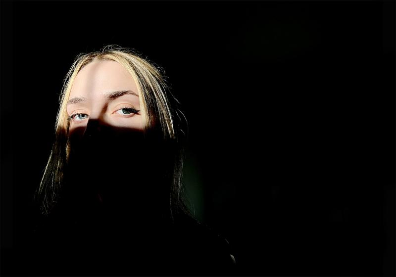 A blonde woman's eyes and head are illuminated by sunlight in an otherwise dark black room.