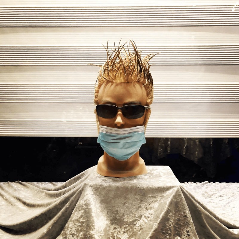 a male mannequin head in a shopwindow wearing sunglasses and a medical mask, but the mask is not covering his nose at all