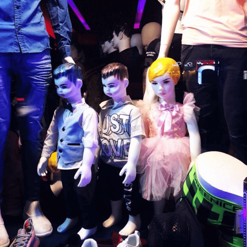 three desperate looking mannequin children standing in a shop window, surrounded by male mannequins in the background, night with neon lights