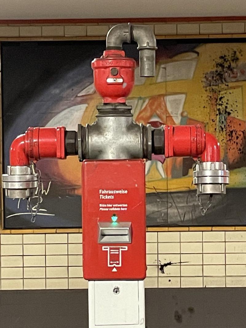 das bild zeigt einen roten fahrkartenentwerter in einer berliner u-bahnstation. direkt dahinter befindet sich ein roter hydrant mit einem anschluss oberhalb und zwei symmetrisch angeordnete, nach unten abgehenden anschlüssen.  