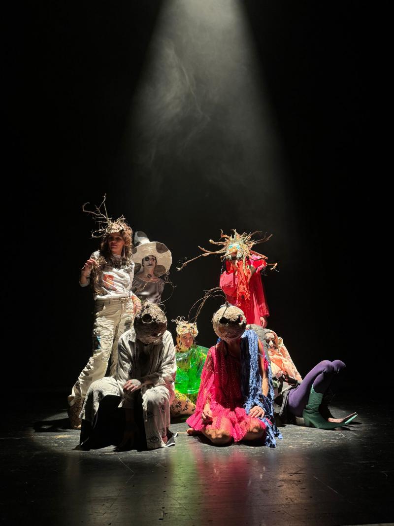 Sieben menschliche, maskierte Figuren in abstrakter Verkleidung stehen dicht aneinander in einem Lichtkegel auf einer dunklen Bühne. 