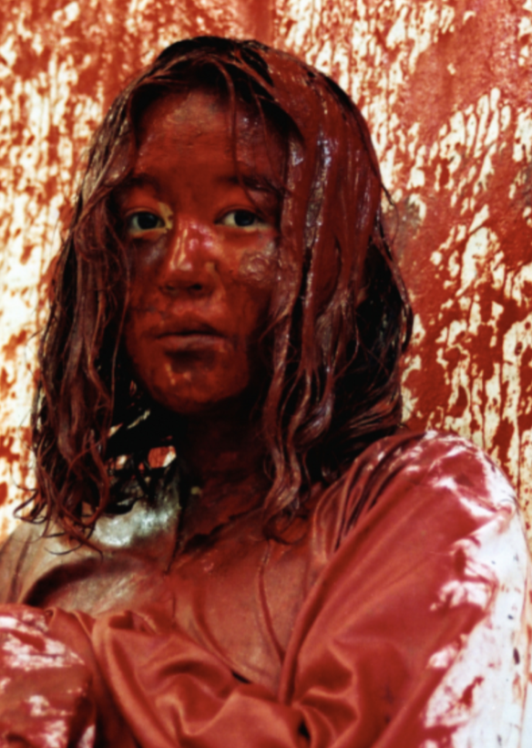 Image of a woman looking at the camera covered in red paint ; both her clothes and the wall are as well splashed with paint 