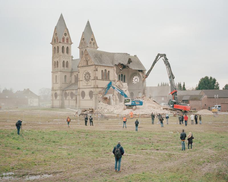 Zwei Bagger reißen eine alte Kirche ab. Hinter einem Bauzaun beobachten Menschen die Szene. 