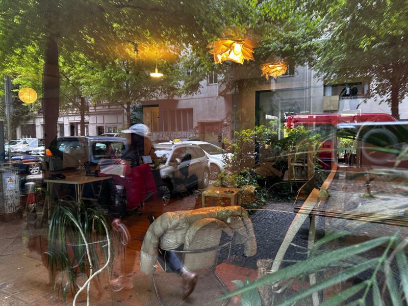 Fotografiert durch ein großes Fenster. Die Straße und eine vorbereigehende Person reflektieren sich im Fenster. Dahinter stehen Pflanzen, Sessel und 2 Deckenlampen.