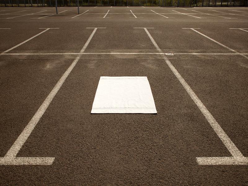 Ein leerer, grau asphaltierter Autoparkplatz mit mehren Parkbuchten, auf der mittleren liegt ein weissen Handtuch.