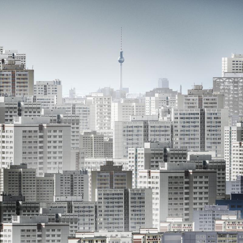 Die Fotocollage zeigt die Skyline des Berliner Bezirks Marzahn-Hellersdorf. Dicht an dicht stehen zahlreiche Plattenbauten mit ihren charakteristischen, einheitlichen Fassaden, die eine fast endlose urbane Struktur ergeben. Die Gebäude variieren in Höhe und Architektur leicht, bleiben aber in ihrer gleichförmigen Erscheinung bestehen. In der Ferne ragt der Berliner Fernsehturm aus der dunstigen Skyline heraus, ein Symbol der Stadt, das in Kontrast zur dichten Wohnbebauung des Vordergrunds steht.