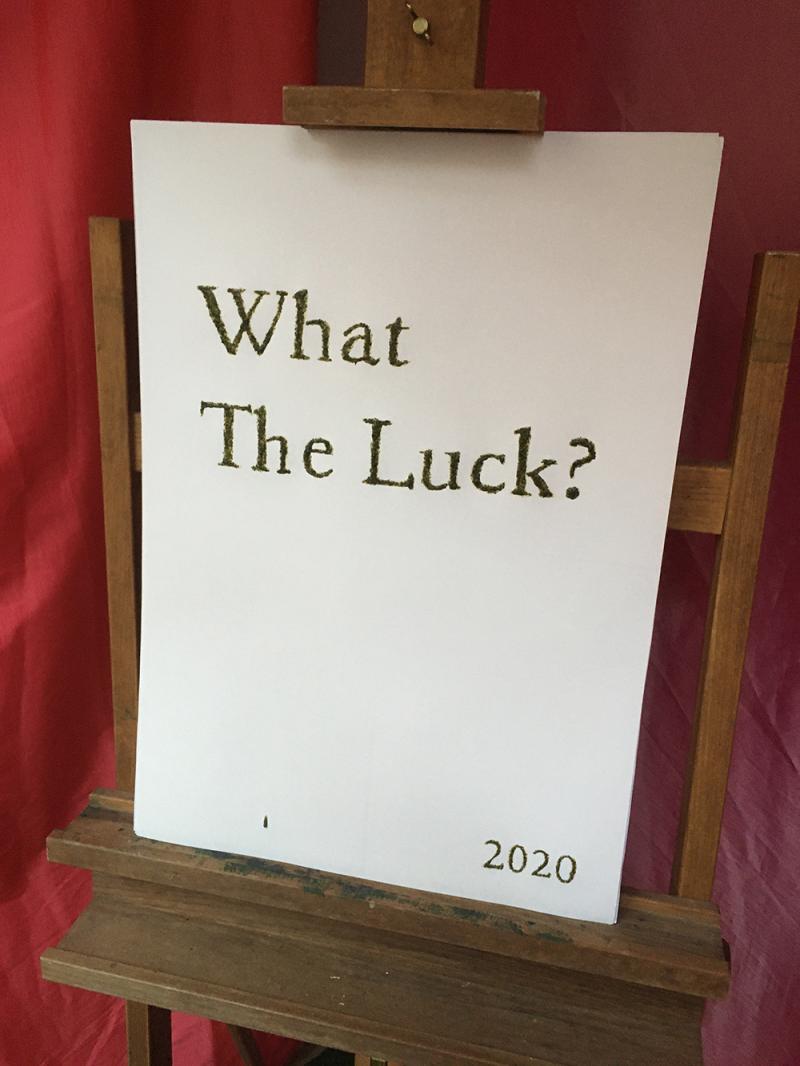 Auf einer Staffelei steht ein A2 Block auf dem der Schriftzug "What The Luck? 2020" steht. Der Schriftzug ist gezeichnet mit einer Tempera aus gemahlenen Kleeblättern 