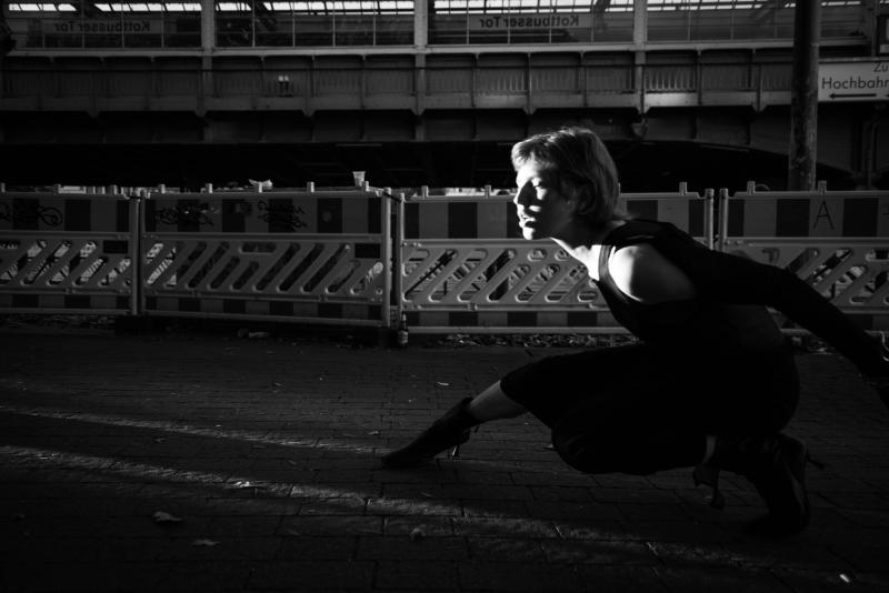 Das Bild zeigt eine Frau in einer dynamischen Pose auf einer urbanen Straße in Schwarz-Weiß. Sie ist halb geduckt, mit einem nach vorne gerichteten Blick und wird von einem dramatischen Lichtstreifen hervorgehoben, der ihr Gesicht betont.
