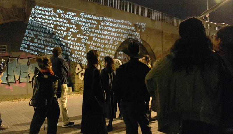 A nighttime scene of participants gathered, watching a projection of text referencing classism, racism, sexism, and other forms of discrimination. This is part of the Gwisin (Ghost) Tour, which explores hidden narratives and marginalized voices in urban spaces.