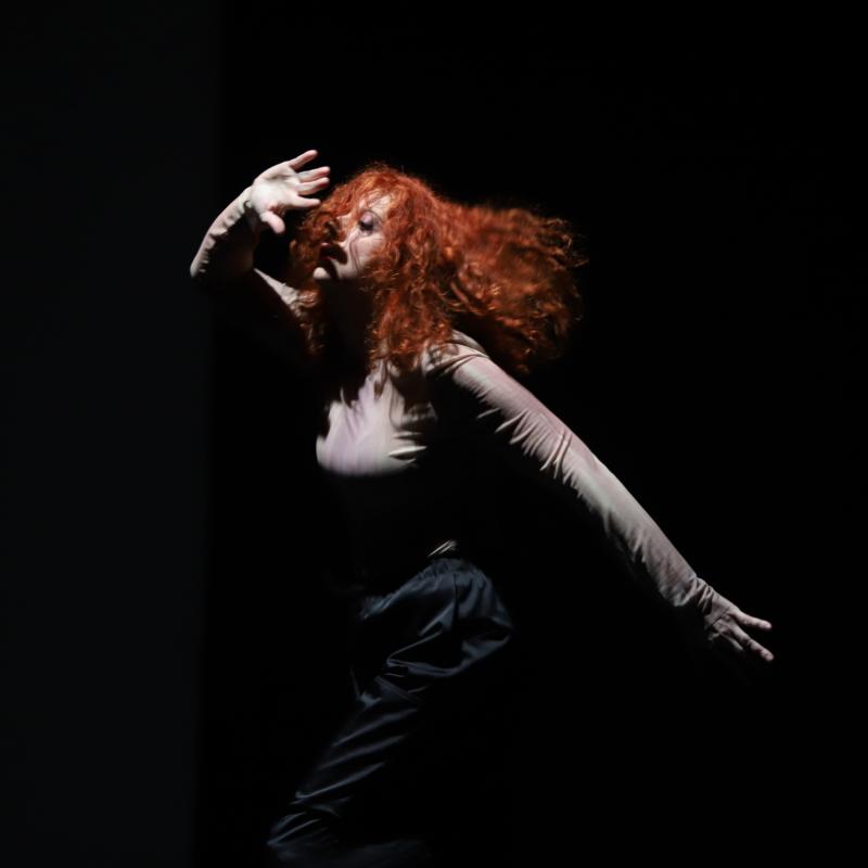 Das Bild zeigt eine Tänzerin mit leuchtend roten, lockigen Haaren, die sich in einer dynamischen Pose befindet. Sie trägt ein langärmeliges, eng anliegendes Oberteil und dunkle Hosen. Ihr Gesicht ist teilweise durch ihr bewegtes Haar verdeckt. Die Beleuchtung hebt sie dramatisch aus einem dunklen Hintergrund hervor, wodurch eine intensive Atmosphäre entsteht.