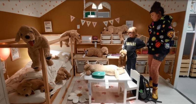 Filmstill. Eine Frau und ein Kind stehen in einem Ikea-Modellschlafzimmer und schauen einem Spielzeughund zu