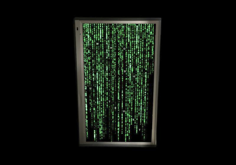 Auf einer Flächenleuchte ein Programmiercode in grün auf schwarzem Untergrund moniert. Einzelne Buchstaben treten besonders hell hervor; diese ergeben den Satz  WHO DO U TRUST. 