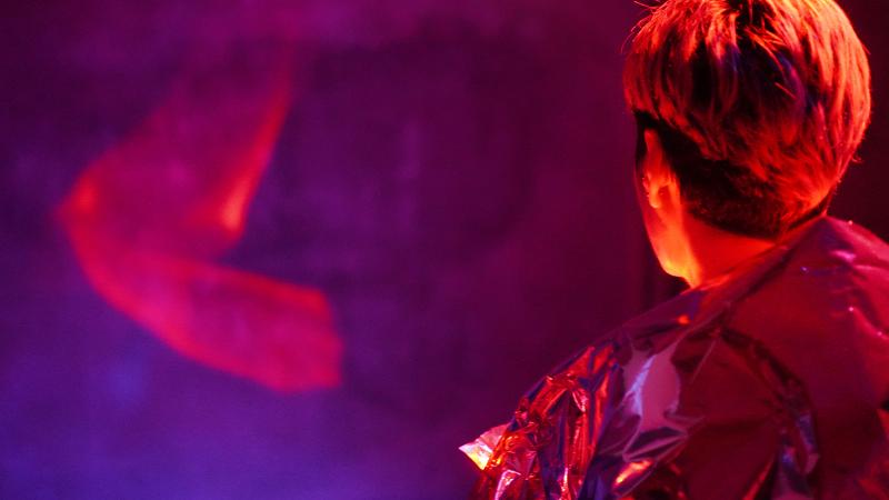 Die performende Person trägt ein reflektierendes Material über den Schultern. Die Person steht mit dem Rücken zu uns vor einer Wand. Das Licht ist blau rot, und es reflektiert auf das Material, die Haare, die Haut und die Wand. Eine rote Reflektion auf der Wand ergibt ein geschwungenes leuchtendes Muster. Es sieht aus wie leuchtender Rauch der schwebt.