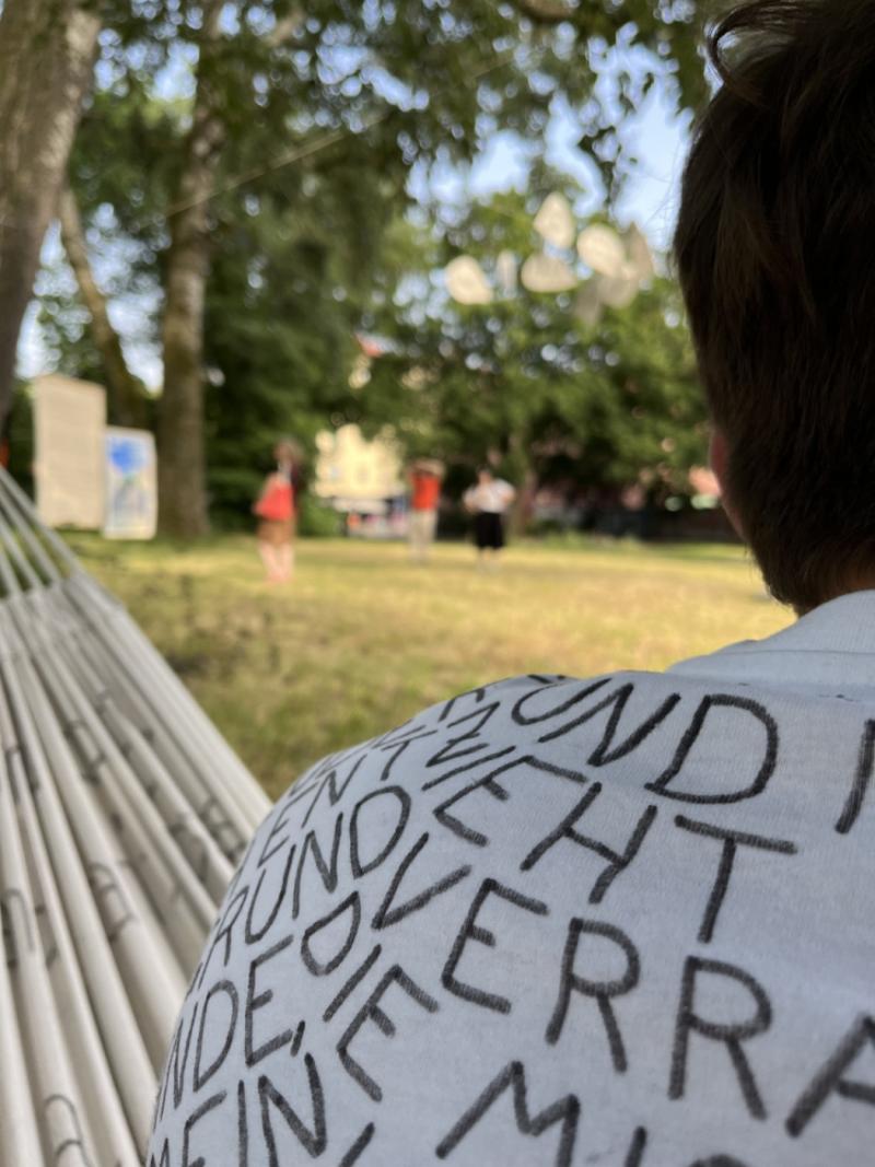 Zu sehen ist die linke Schulter einer Person, die auf einer Hängematte sitzt. T-Shirt und Hängematte sind weiß und mit Marker beschriftet. Im Hintergrund ist ein Park.