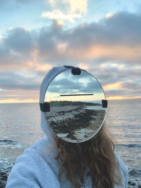 Frau mit Spiegel vor dem Gesicht. Im Hintergrund ist das Meer und der Himmel zu sehen.