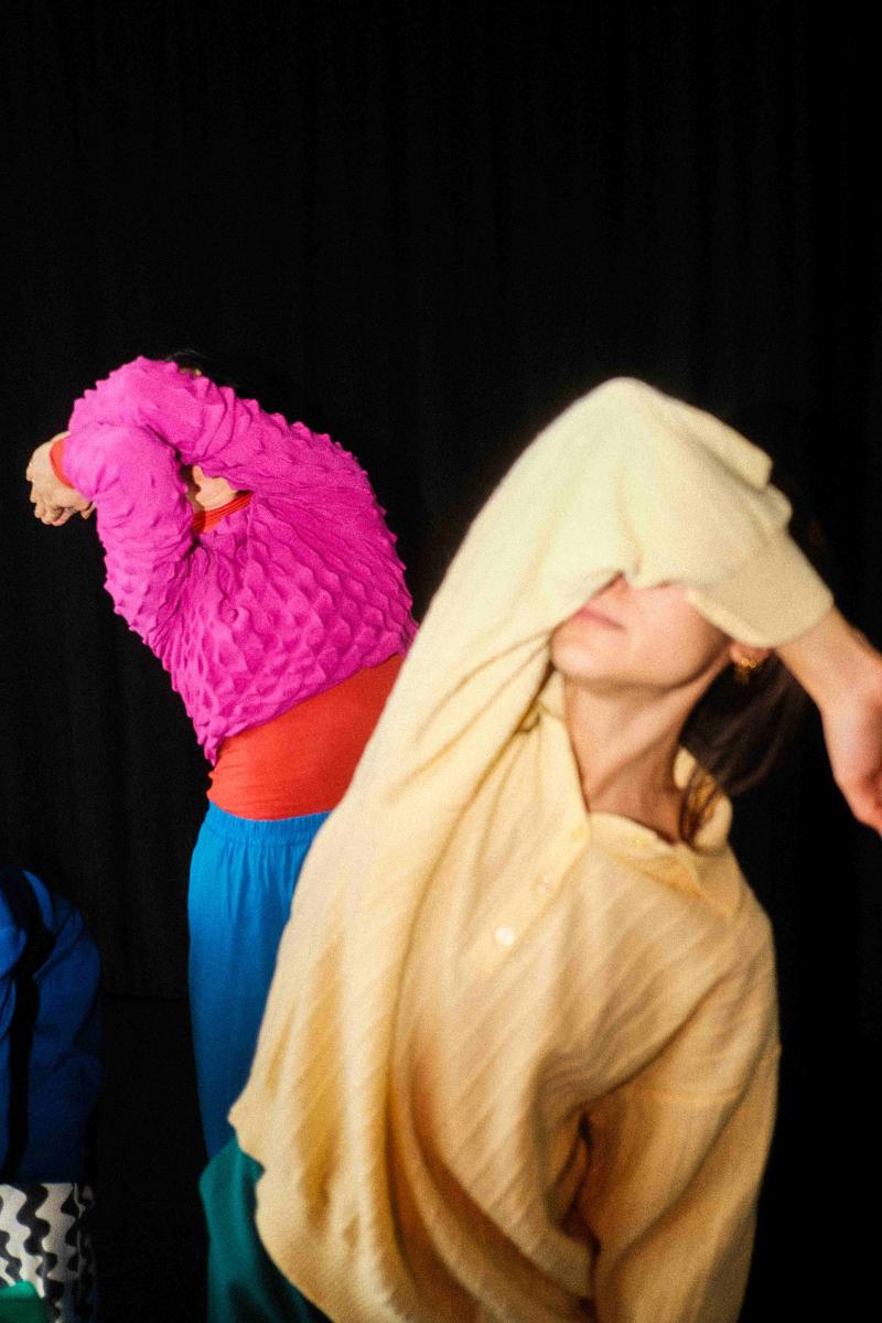Two dancers in mid-motion against a dark background, partially obscuring their faces with their arms. The dancer in the foreground wears a pale yellow oversized shirt, their arm covering their eyes as they tilt their head. Behind them, another dancer in a textured magenta top and blue pants mirrors the movement, their arms forming a dynamic shape. The composition captures fluidity and abstraction, emphasizing texture, color, and form.