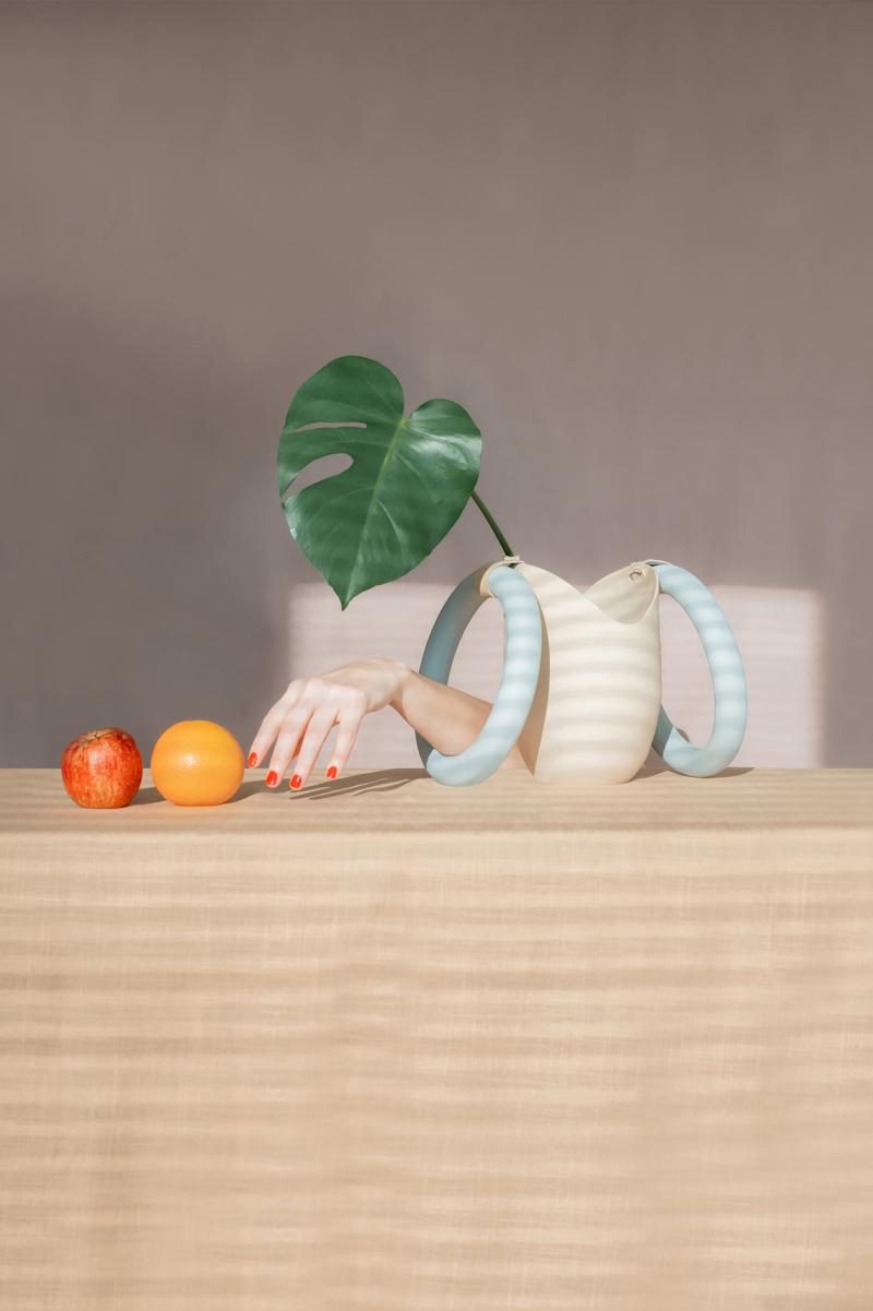 Ein Apfel, eine Orange, eine Frauenhand und eine Vase mit einem Monsterablatt auf einem Tisch