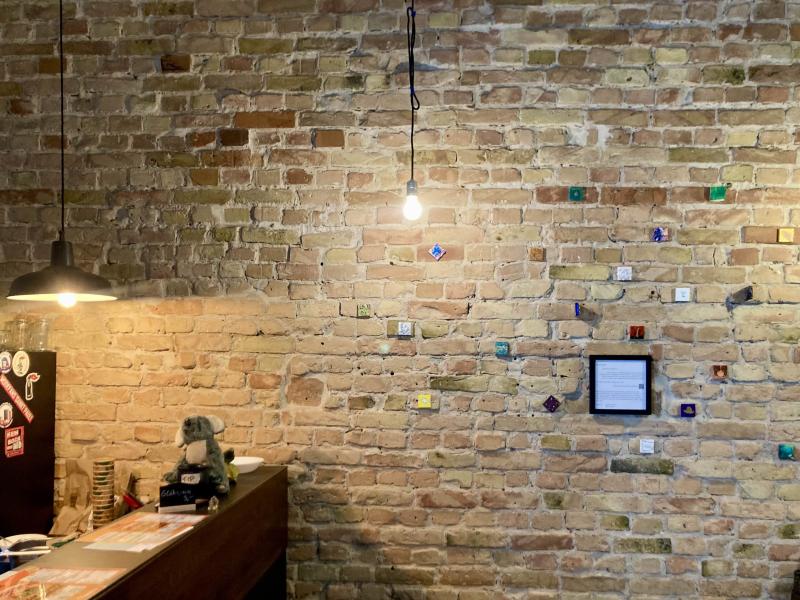 Auf einer Backsteinmauer in einem gemütlich beleuchteten Innenraum hängen viele bunte Bilder, die alle ganz klein sind. 