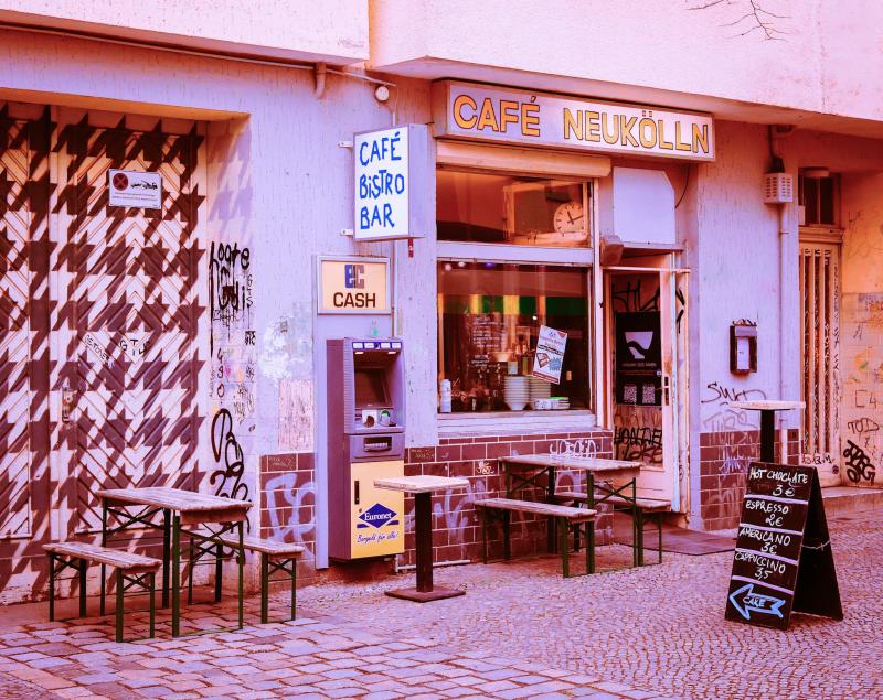 Vor einem Garagentor stehen Stühle und Bänke, an der Wand daneben ein Geldautomat, viele Berliner Grafitti. Links davon das Café wirkt einladend und trägt die Überschrift "Café Neukölln". Im Schaufenster sind Kuchen und Cocktails zu sehen. Und natürlich ein Lächeln.  Stelle Dir vor, Du warst noch nie dort, fühlst Dich aber sofort wie zuhause. 