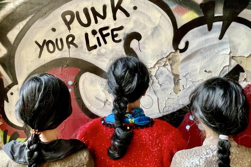 3 Personen mit schwarzen Zöpfen stehen mit dem Rücken zu uns und blicken auf eine Grafffitiwand. Auf der Wand steht "Punk Your Life".