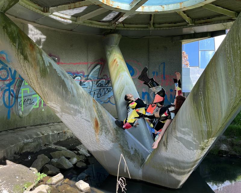zwei Tänzerinnen in bunten Kostümen zwischen Brückenpfeilern vor einer Wand mit Graffiti