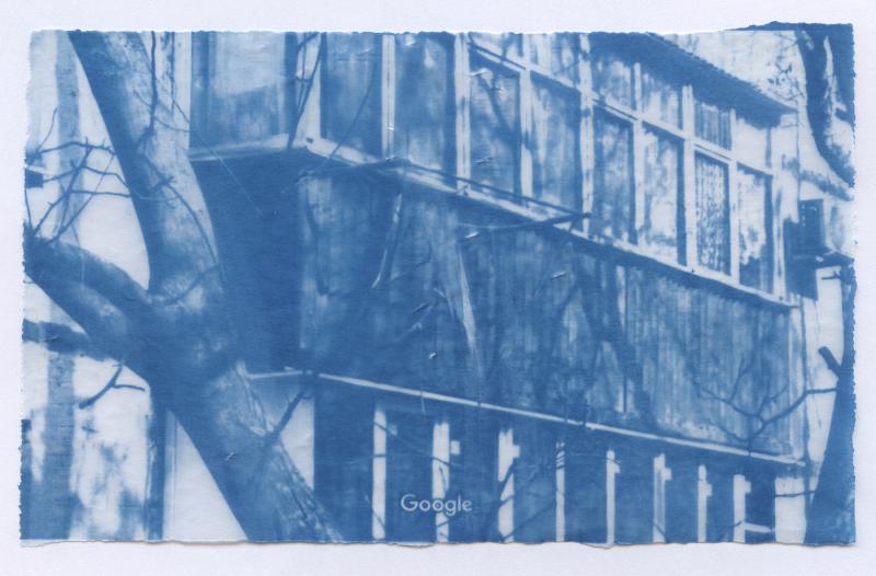 A cyanotype print on tracing paper depicting a close-up view of a weathered residential building, captured from a Google Street View screenshot. The composition is dominated by a balcony of a post-Soviet panel apartment block from the 1980s, its worn facade partially obscured by bare tree branches in the foreground. The Google watermark is visible at the bottom. Rendered in deep blue tones characteristic of the cyanotype process.
