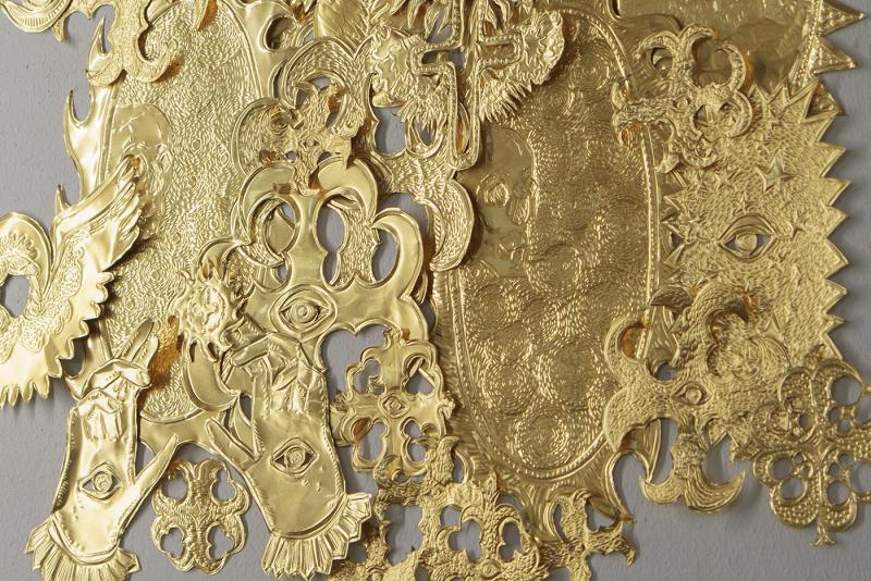 The installation is composed of hand-crafted Milagros: eyes, angels, crosses, and other protective symbols. They are arranged above a dense formation of sheep that are gathered tightly, almost as a single body.
