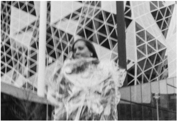 A grainy, black and white photograph showing a blurred figure of a person in the foreground, seemingly draped in translucent or reflective material. In the background, a large modern building with a complex, geometric triangular facade creates a sharp contrast with the fluid, out-of-focus movement of the protagonist.