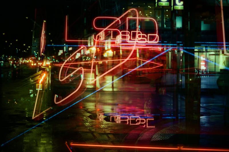 Das mehrfach belichtete Bild zeigt eine Straße mit Neonlichtern mit dem Schriftzug „LSD". Es ist dunkel, und die Straße ist aufgrund von Regen nass; im nassen Asphalt spiegelt sich das Neonlicht.